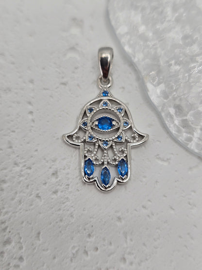 Embrace the protective energies of the Hamsa Hand with this exquisite pendant, meticulously crafted from 925 sterling silver. At its center lies a captivating blue evil eye crystal, symbolizing vigilance against negative energies and a beacon of good fortune.

The pendant's intricate detailing is further enhanced by additional blue crystals, gracefully accentuating its contours and adding a touch of elegance. Each crystal is precisely set, amplifying the protective gaze of the central motif and highlighting
