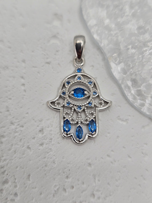 Embrace the protective energies of the Hamsa Hand with this exquisite pendant, meticulously crafted from 925 sterling silver. At its center lies a captivating blue evil eye crystal, symbolizing vigilance against negative energies and a beacon of good fortune.

The pendant's intricate detailing is further enhanced by additional blue crystals, gracefully accentuating its contours and adding a touch of elegance. Each crystal is precisely set, amplifying the protective gaze of the central motif and highlighting
