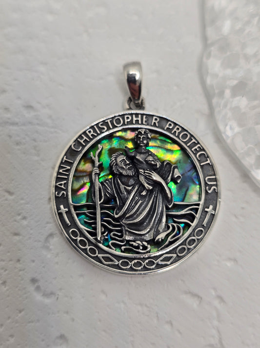 This Saint Christopher Pendant blends spiritual symbolism with striking natural beauty. Crafted from solid 925 sterling silver, the pendant features a luminous abalone shell inlay, creating a vibrant play of colors behind the detailed image of Saint Christopher, the patron saint of travelers.

Surrounding the central figure are the words: "Saint Christopher Protect Us"—a heartfelt prayer carried through generations by those seeking safe passage and divine guidance. The saint is shown mid-journey, carrying t